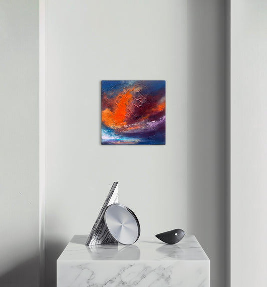A square abstract wall art print with vibrant orange, red, blue, and purple brushstrokes, displayed on a light gray wall above a white marble console table with modern silver and black decor.