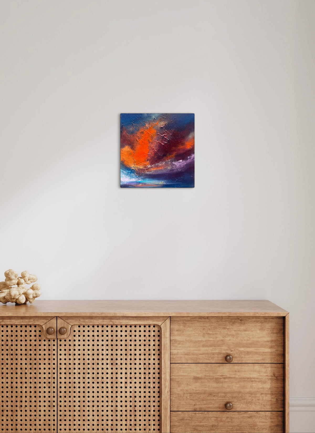 An abstract canvas wall art print featuring vibrant orange, red, and blue brushstrokes, displayed on a white wall above a light wood credenza.