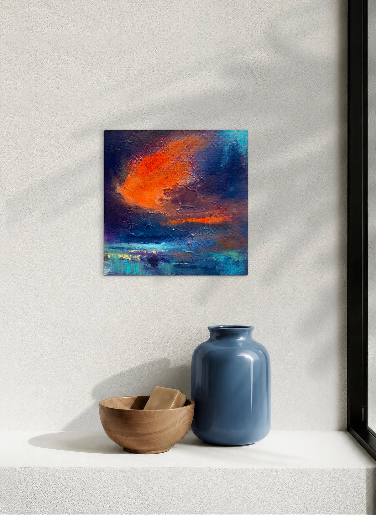 An abstract square canvas painting with vibrant orange, red, and deep blue brushstrokes, displayed on a textured white wall above a white shelf holding a blue ceramic vase and a wooden bowl.