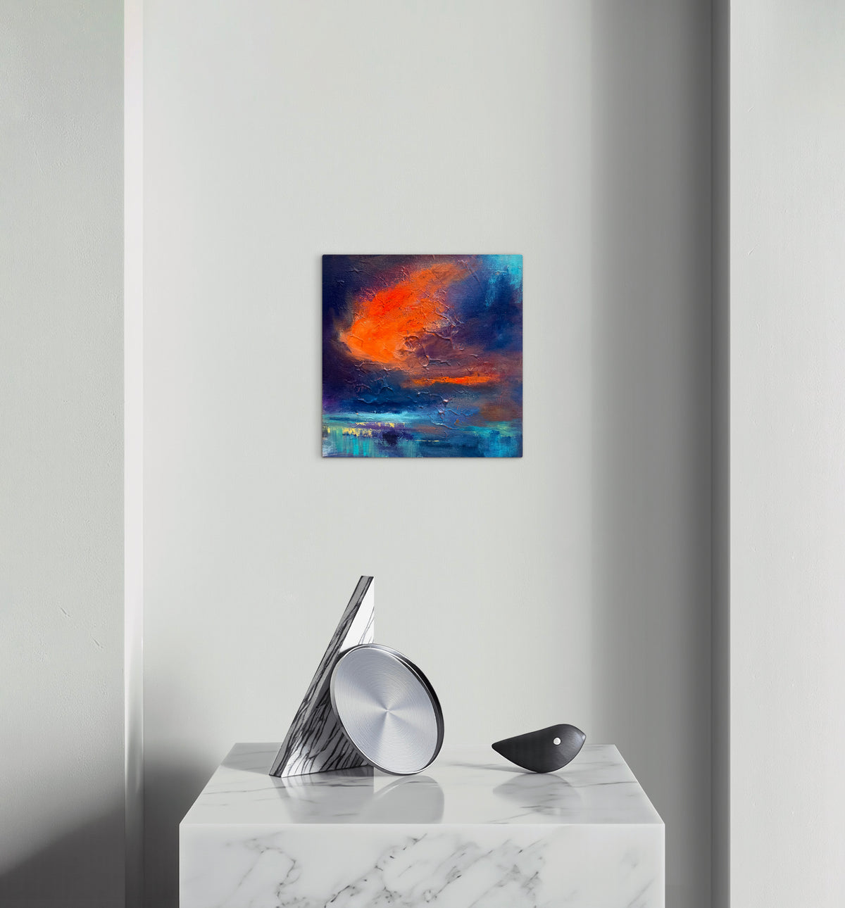 A square abstract canvas print with vibrant orange, red, and deep blue brushstrokes, displayed on a light gray wall above a white marble console table with modern silver and black decor.