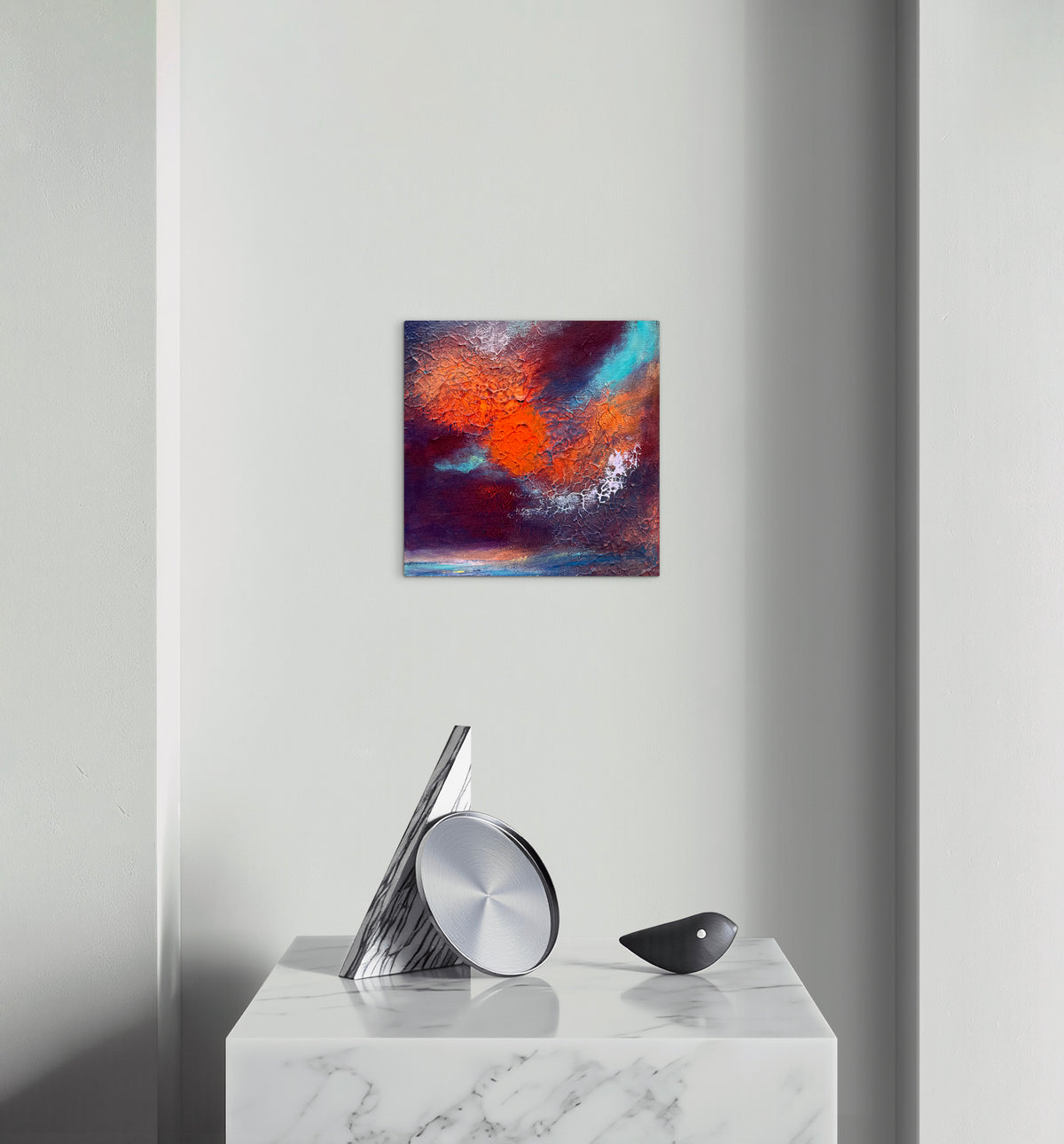 A square abstract canvas print with vibrant orange, red, blue, and purple brushstrokes, displayed on a light gray wall above a white marble console table with modern silver and black decor.