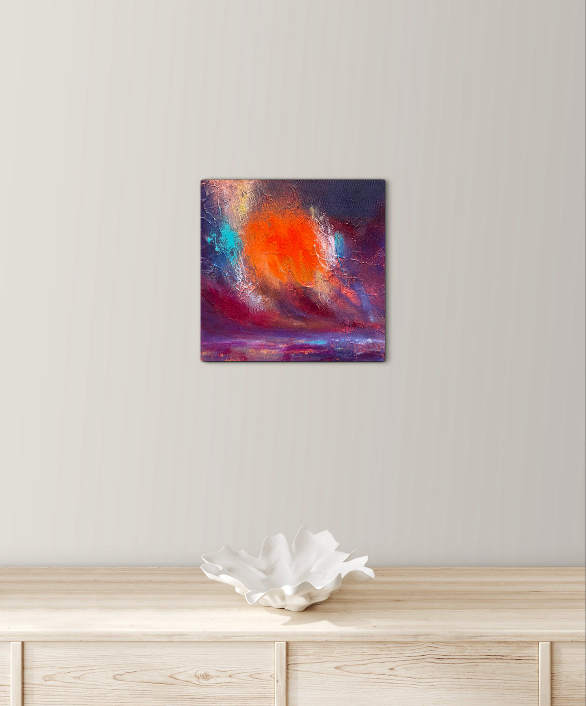 A square abstract wall art print with a vibrant orange sun and purple, blue, and red clouds, displayed on a light beige wall above a wooden console table with a white decorative bowl.