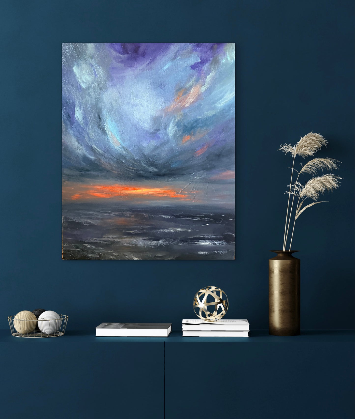 An abstract canvas painting of a dramatic storm sky with purple, blue, gray, and orange clouds, displayed on a dark blue wall above a shelf with decorative books, a vase, and dried grass.