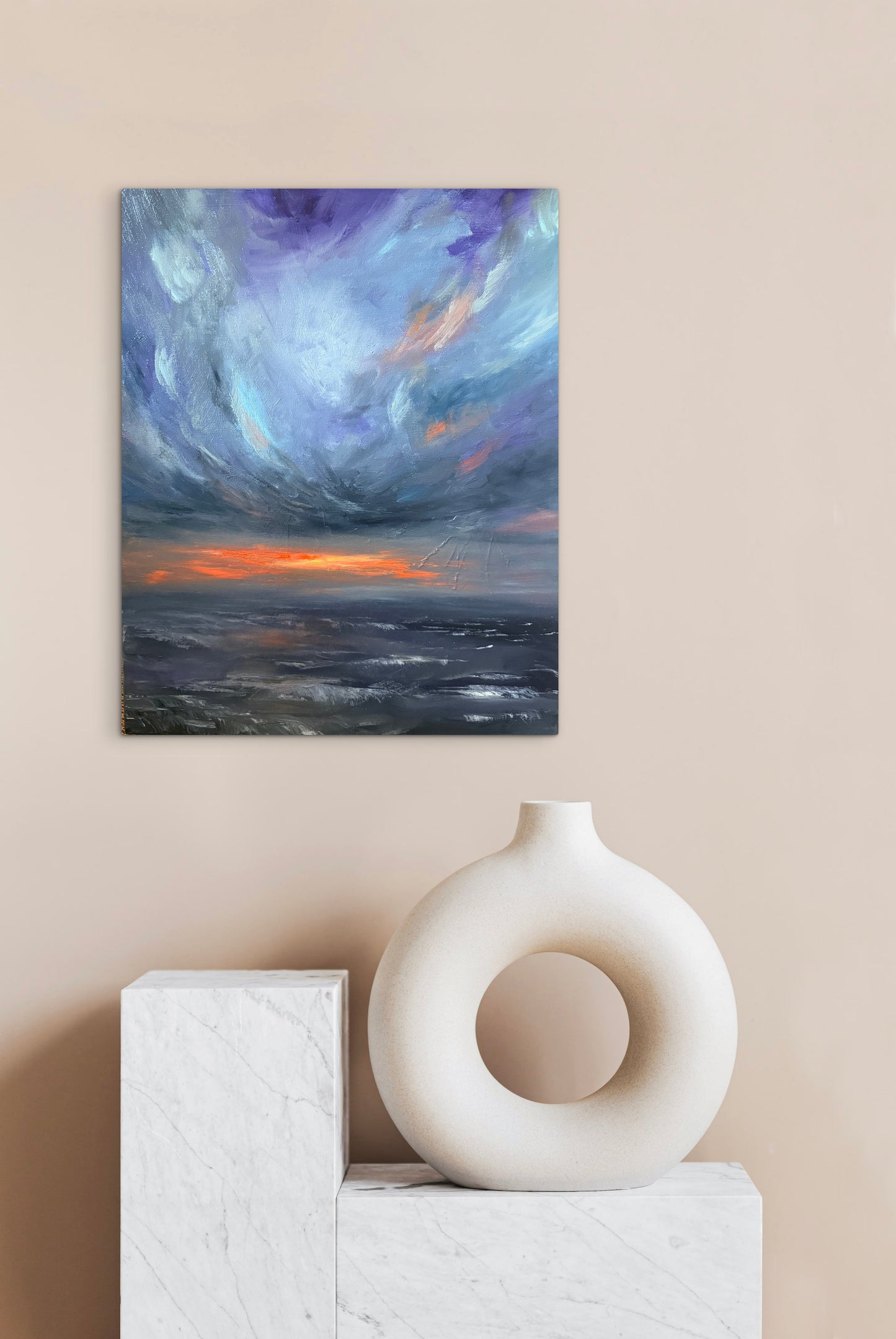 An abstract canvas painting of a dramatic storm sky with purple, blue, and orange tones, displayed on a beige wall above a white marble pedestal with a white ceramic vase.