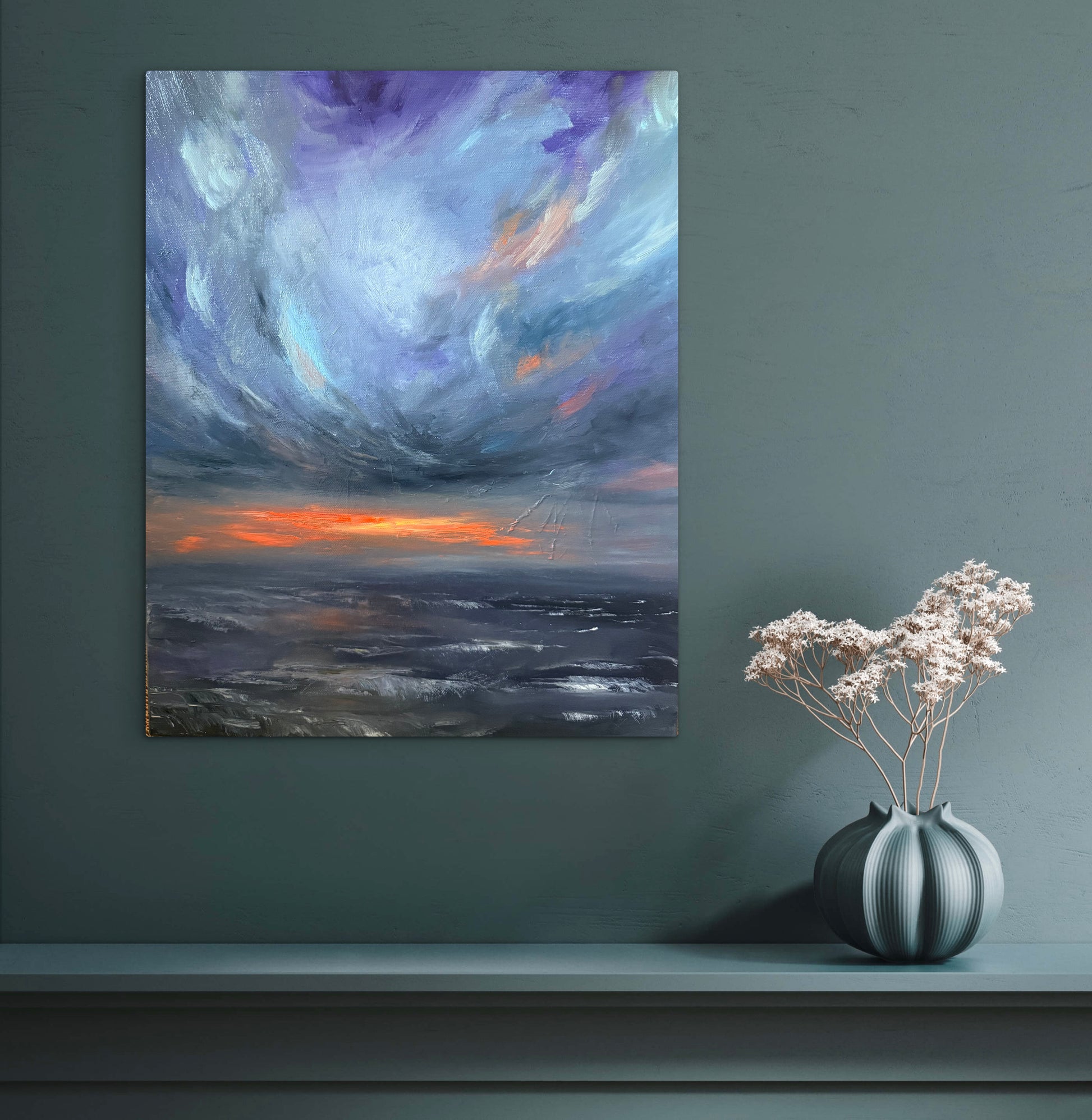 An abstract landscape canvas print featuring a dramatic purple and blue sky over a dark sea with a vibrant orange sunset, displayed on a dark teal wall above a mantel with a gray vase of dried flowers.