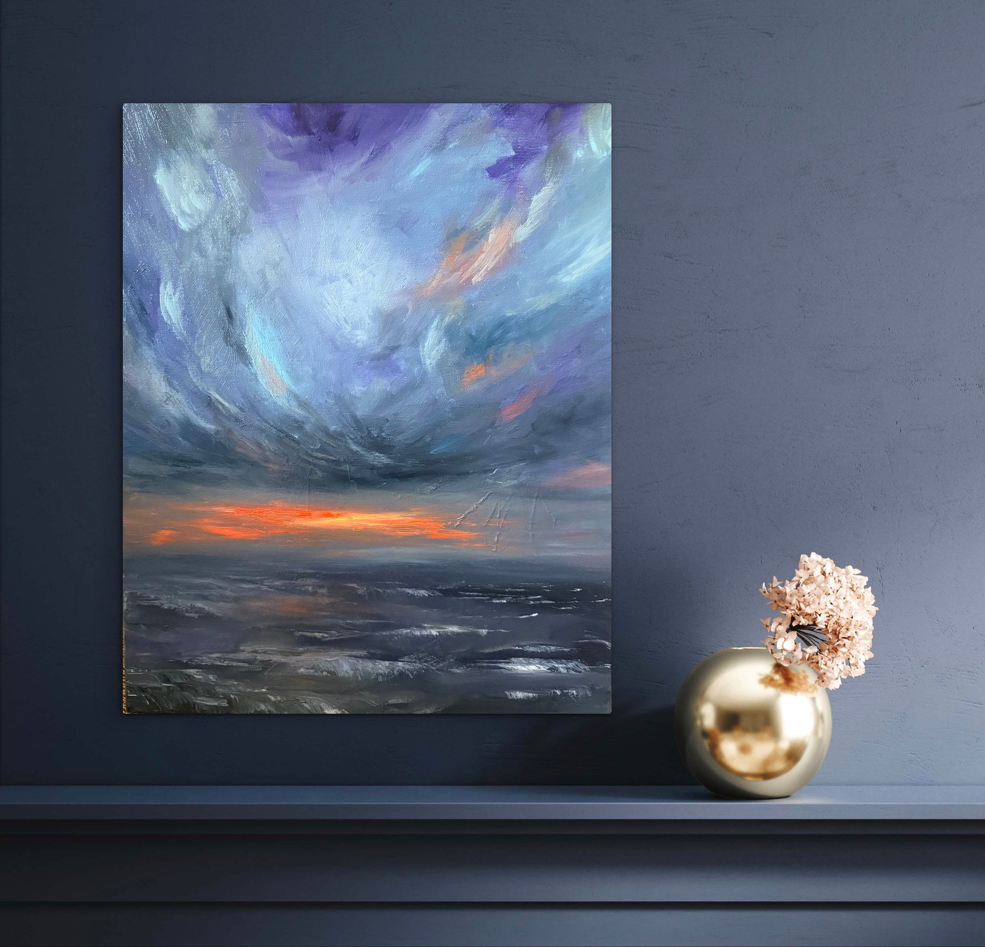An abstract canvas painting of a dramatic storm sky over the ocean, displayed on a dark blue-gray wall above a mantel with a gold vase and dried flowers.