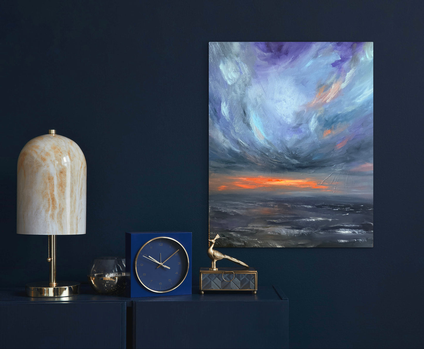 An abstract landscape painting featuring a dramatic stormy sky in shades of purple, blue, and white with a vibrant orange sunset over a dark sea, displayed on a dark navy wall above a console table with a marble lamp, blue clock, and small gold figurine.