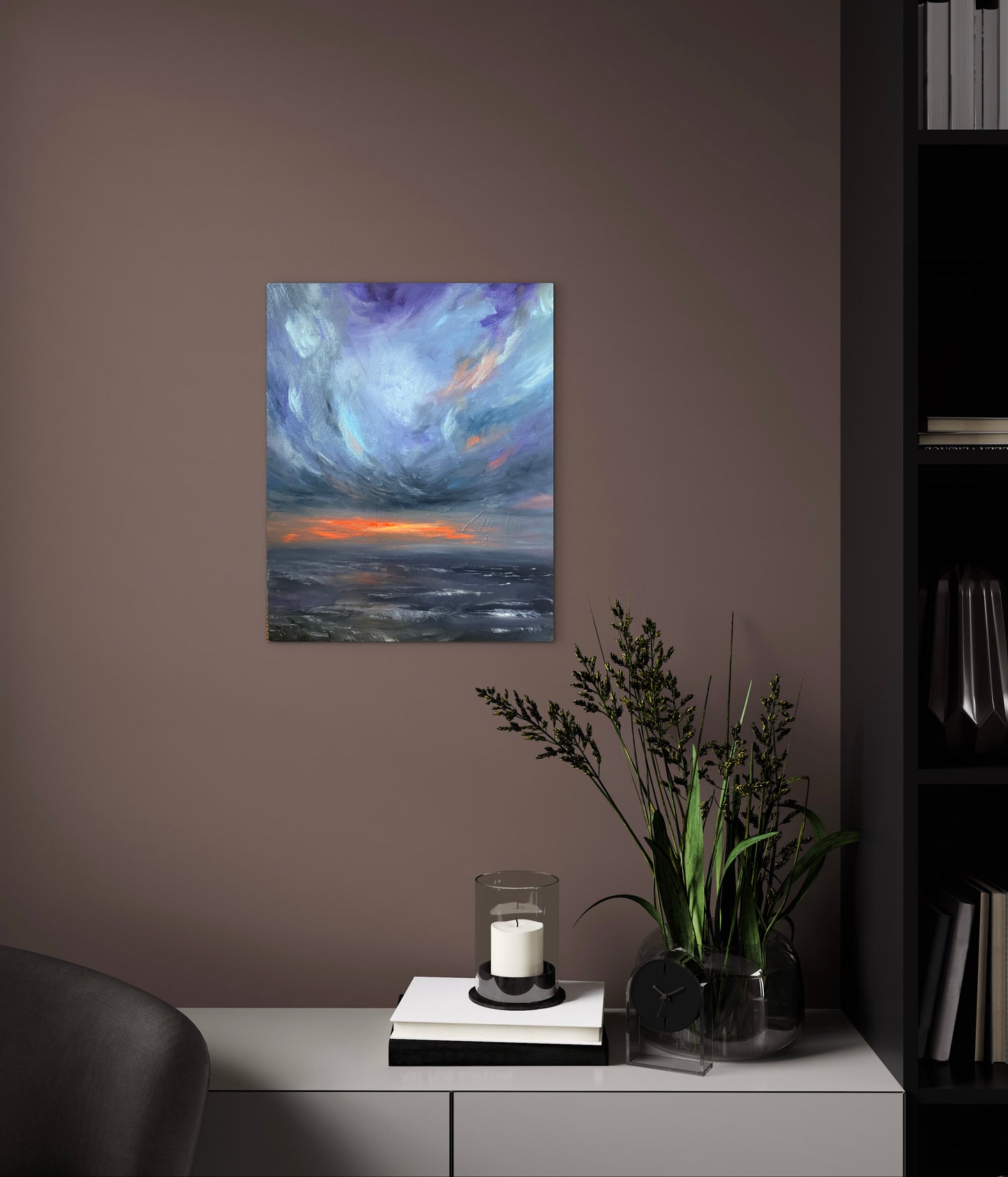 A vertical canvas print of an impressionistic stormy seascape with a glowing orange horizon, displayed on a dark brown wall above a console table with a candle, books, and a potted plant.