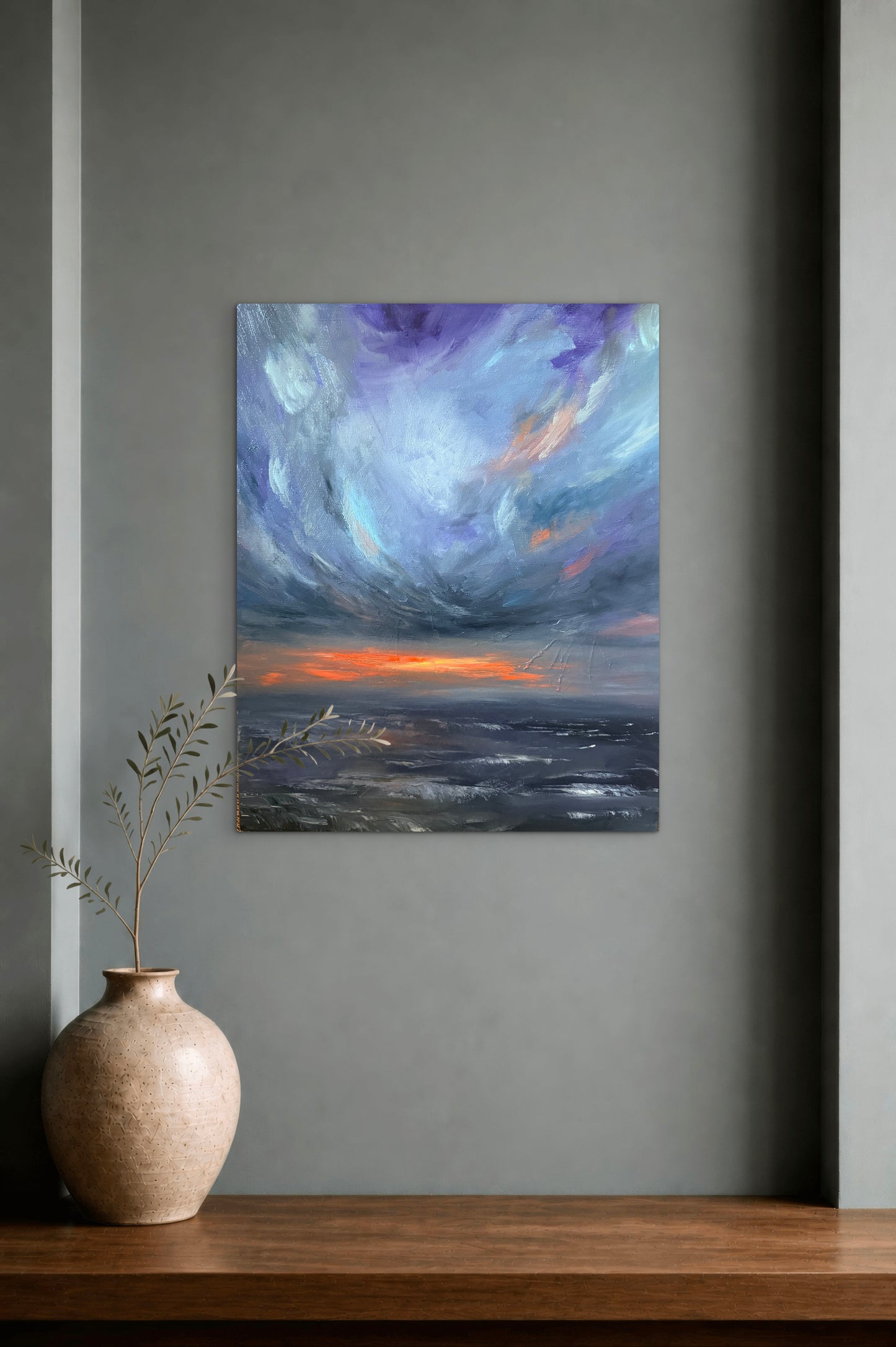 An abstract landscape painting featuring a dramatic purple and blue sky over a dark sea with a bright orange sunset, displayed on a gray wall above a wooden console table with a ceramic vase.