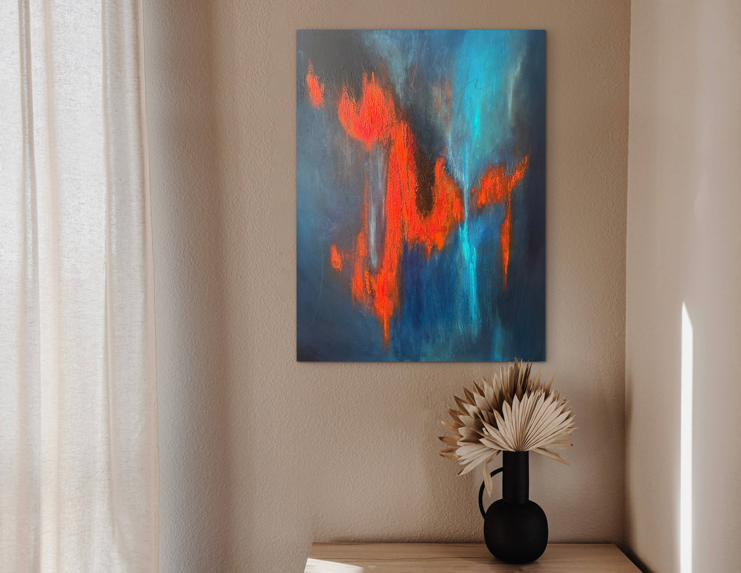 Abstract wall art featuring bold blue and orange brushstrokes, displayed on a beige wall above a small wooden table with a black vase holding dried palm leaves.