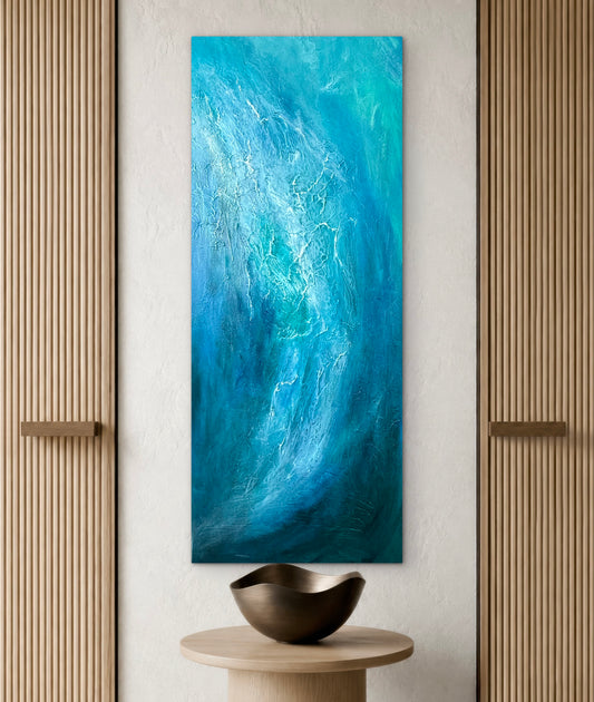 A vertical abstract canvas painting featuring swirling shades of blue and turquoise, displayed on a textured cream wall above a small wooden table with a dark bowl, flanked by ribbed wood panels.