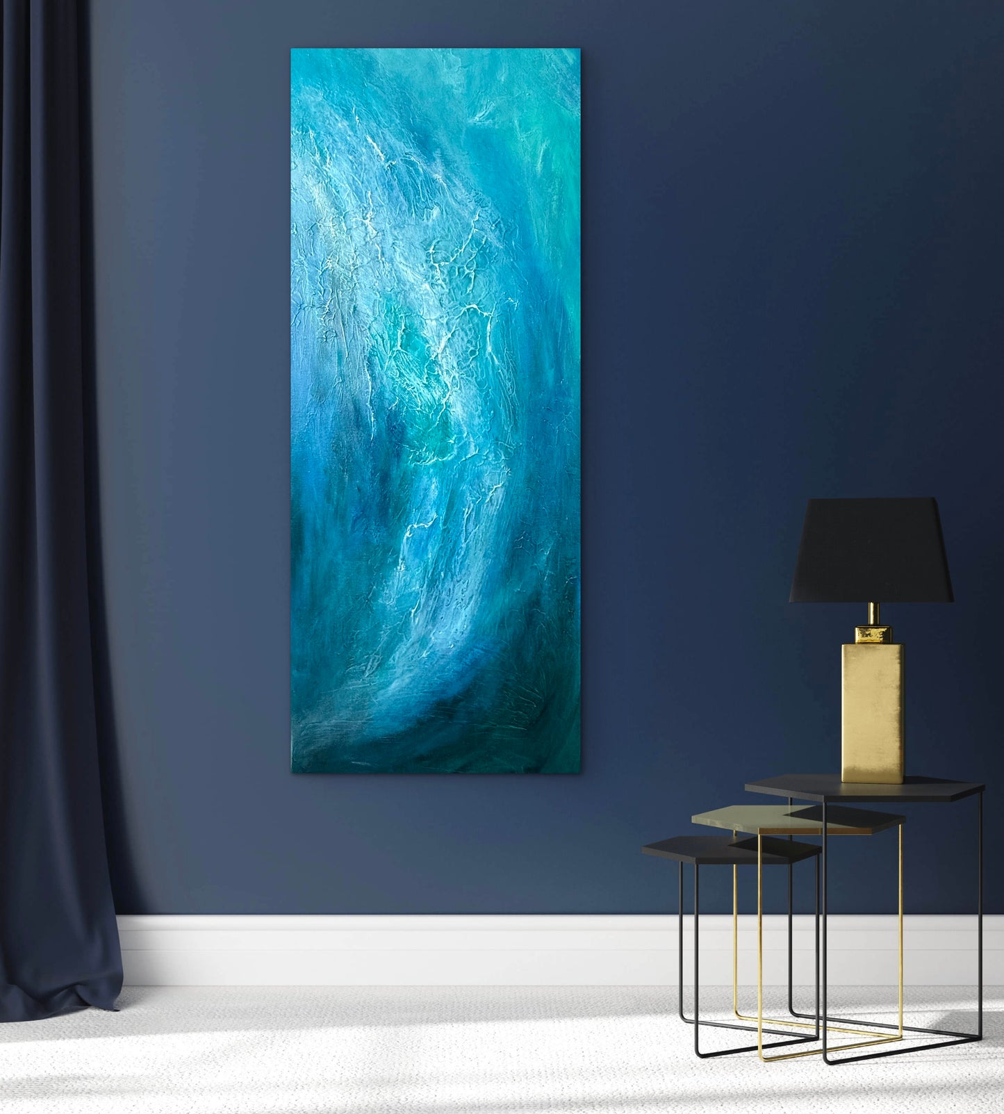 A tall vertical abstract canvas painting with swirling shades of blue, teal, and turquoise, displayed on a dark navy wall in a modern room setting next to a black curtain and a gold table lamp.