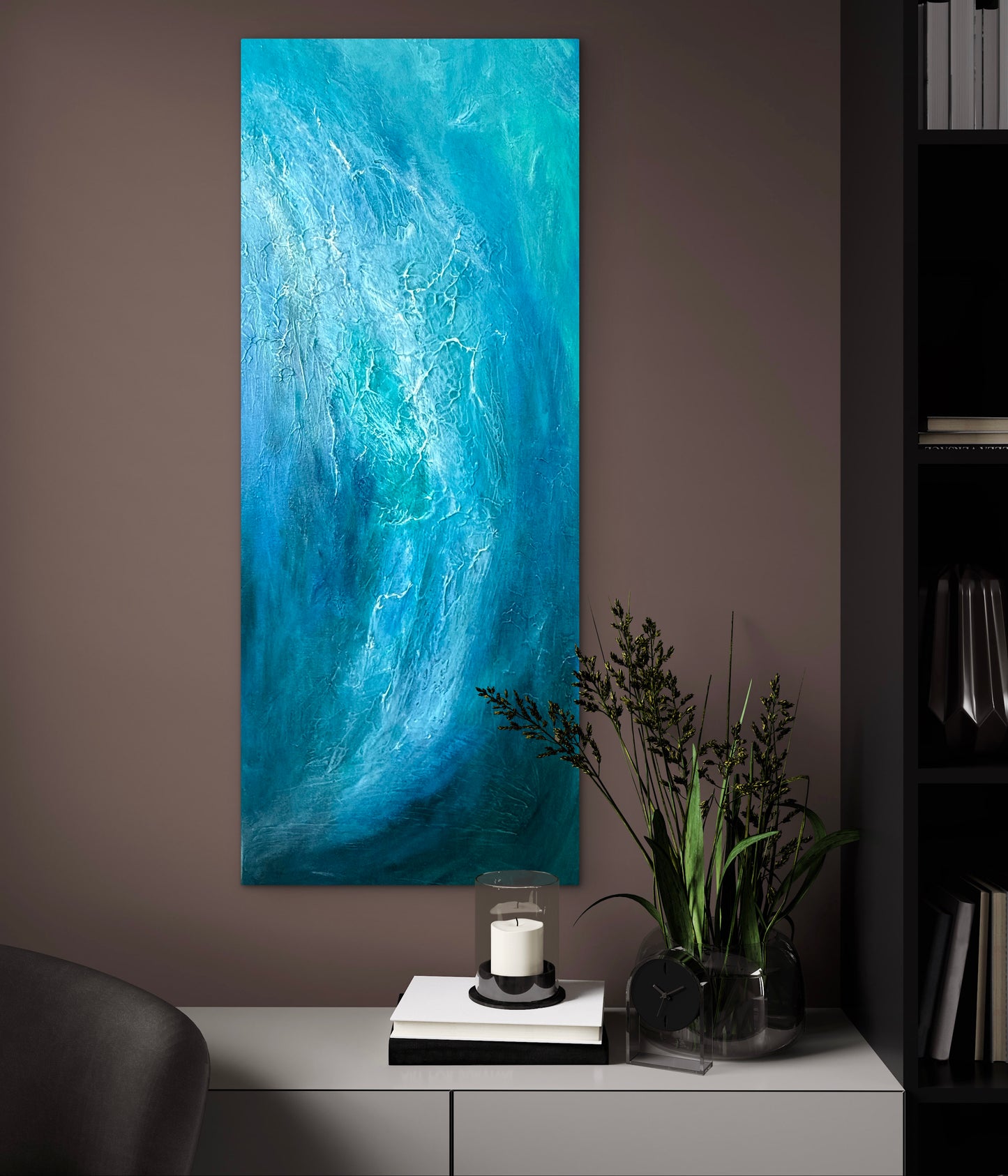 A tall vertical canvas print featuring an abstract blue and turquoise wave design, displayed on a dark brown wall above a console table with a candle, books, and a plant.