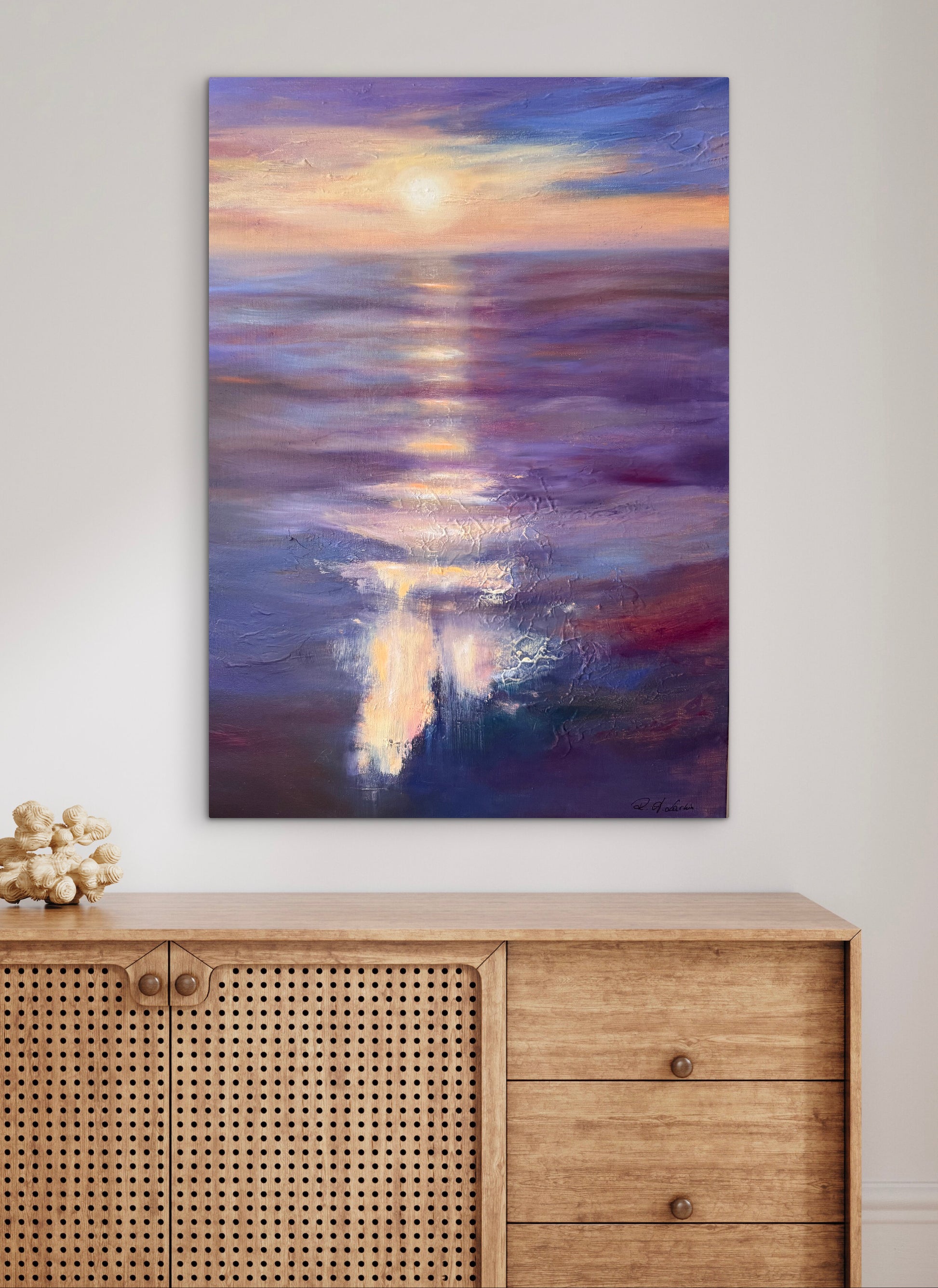 An abstract canvas painting of a sunset over water, featuring purple, blue, and gold hues, displayed on a light gray wall above a wooden cabinet.