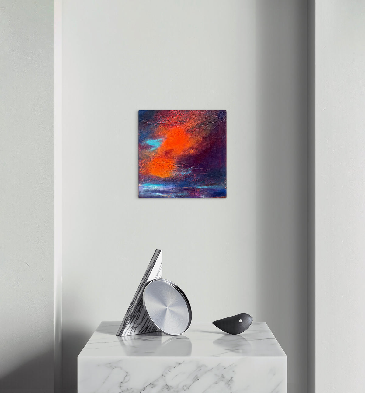 A square abstract wall art print with vibrant orange, red, purple, and blue brushstrokes, displayed on a light gray wall above a white marble console table with modern silver and black decor.