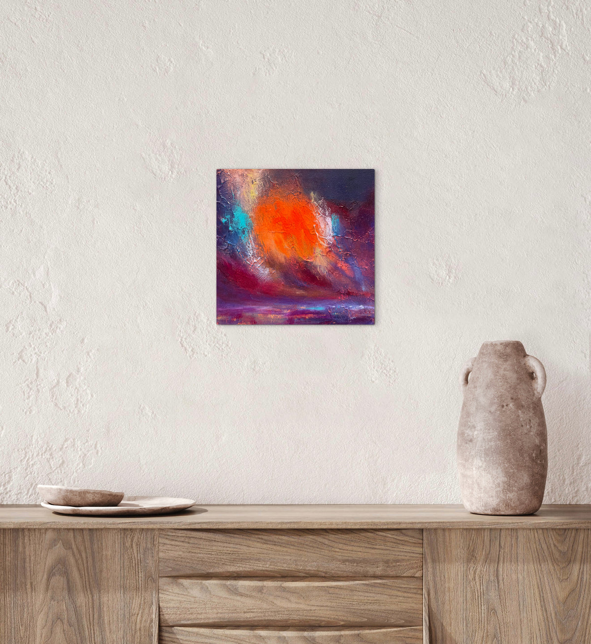 A square abstract wall art print featuring a vibrant orange sun and colorful clouds, displayed on a textured white wall above a wooden sideboard with decorative ceramic objects.