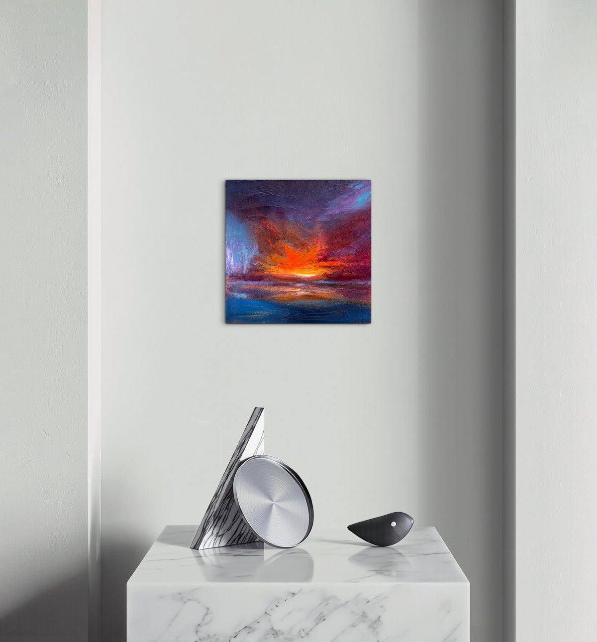 A square abstract canvas print featuring a vibrant sunset or sunrise with deep blue, purple, red, and orange tones, displayed on a light gray wall above a white marble side table with modern silver and black decor.