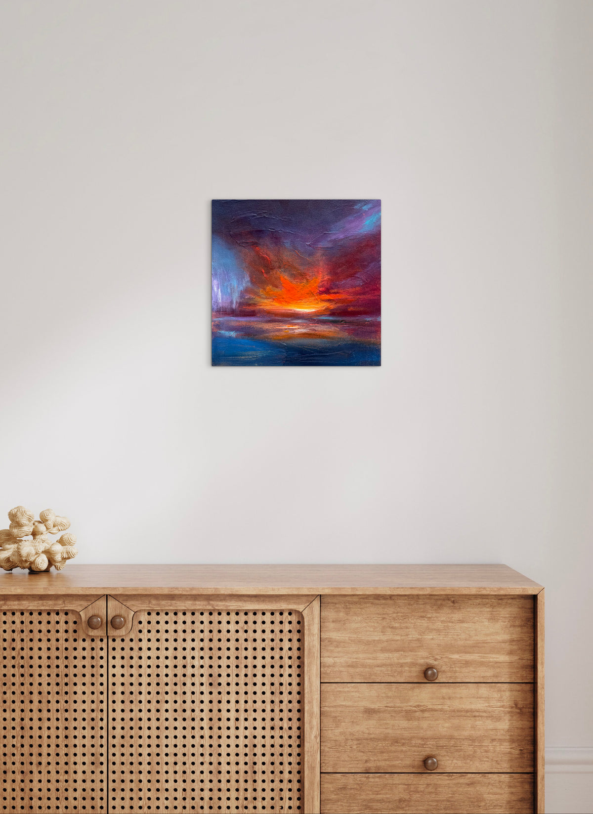 A square abstract canvas print of a sunset over water, featuring vibrant orange, red, and blue hues, displayed on a white wall above a light wood credenza.